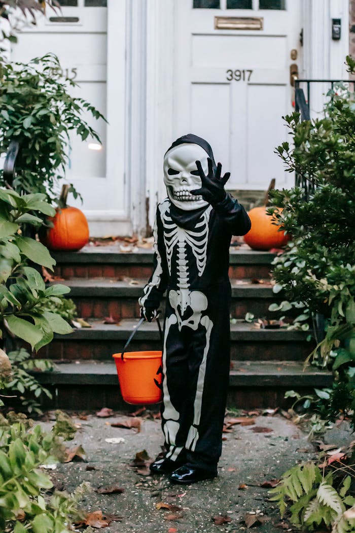 Child in skeleton outfit holding an orange bucket, trick-or-treating on Halloween outdoors.